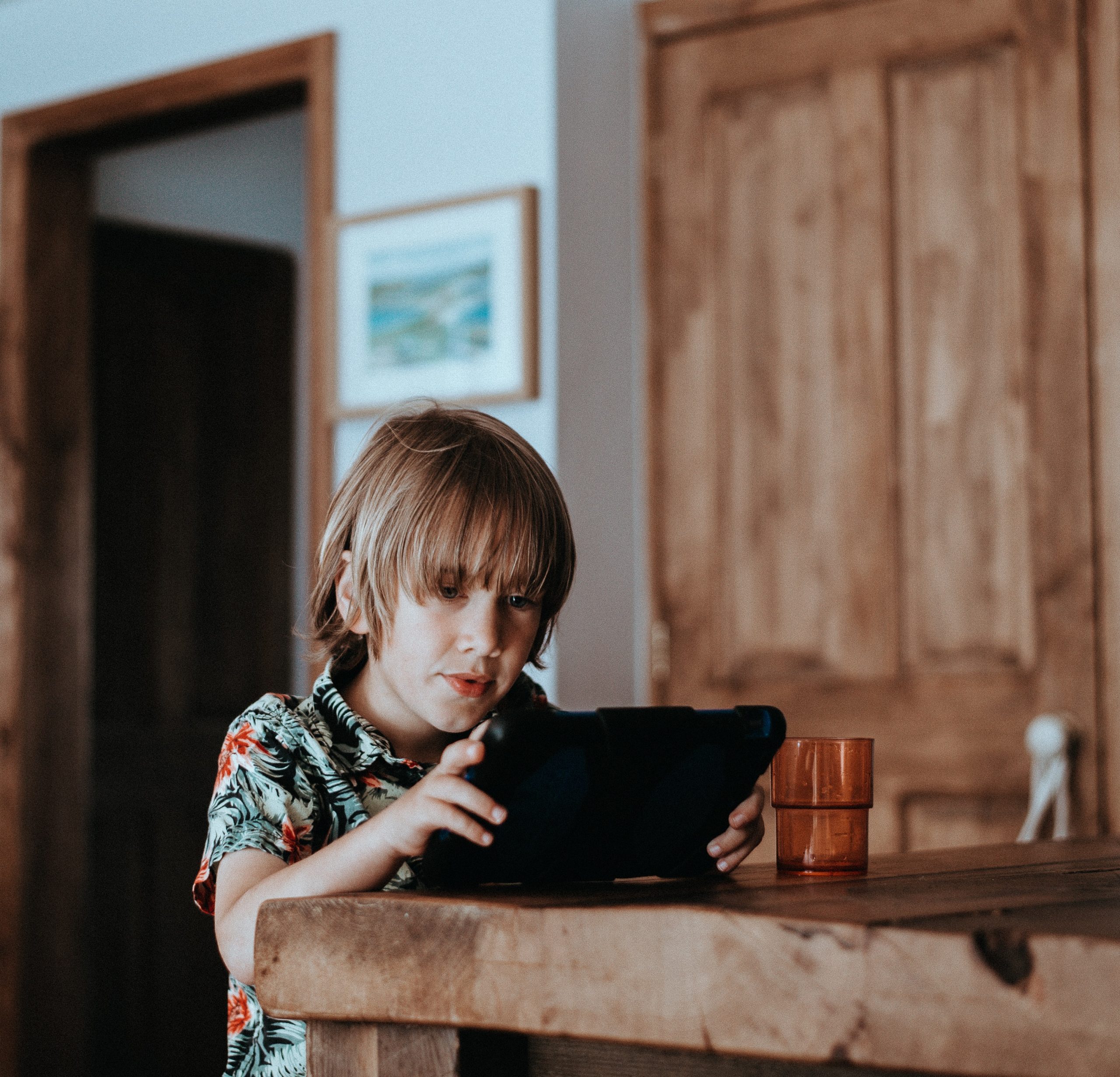 Kind sitz mit eienm Tablet ind er HAnd an einem Holztisch